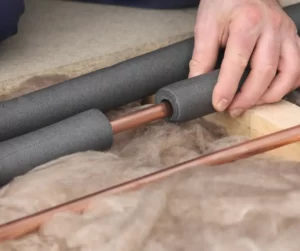 A hand installing foam pipe insulation on copper pipes surrounded by attic insulation.