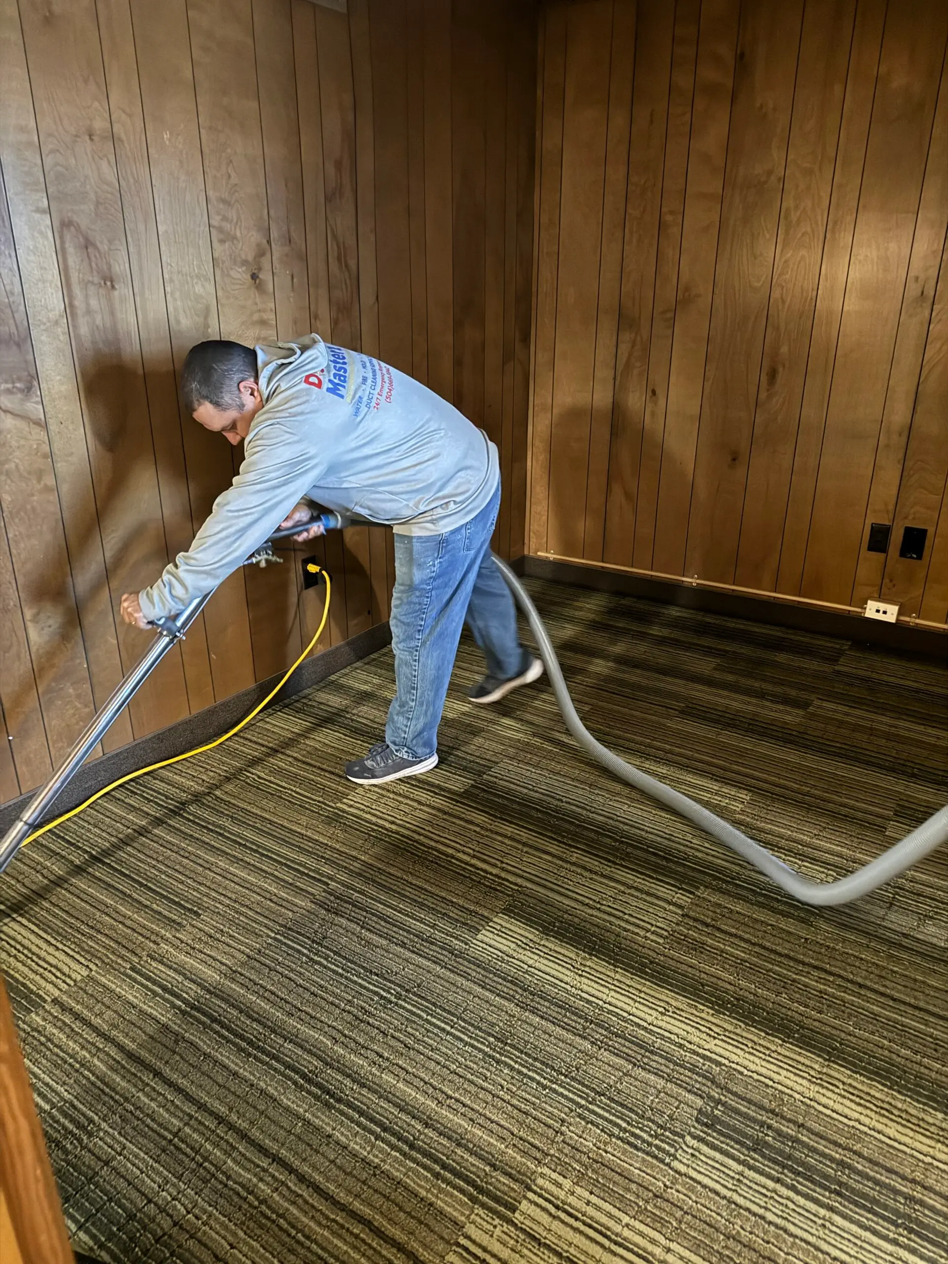 A professional cleaning a wet carpet in a wood-paneled room with a water extraction machine.